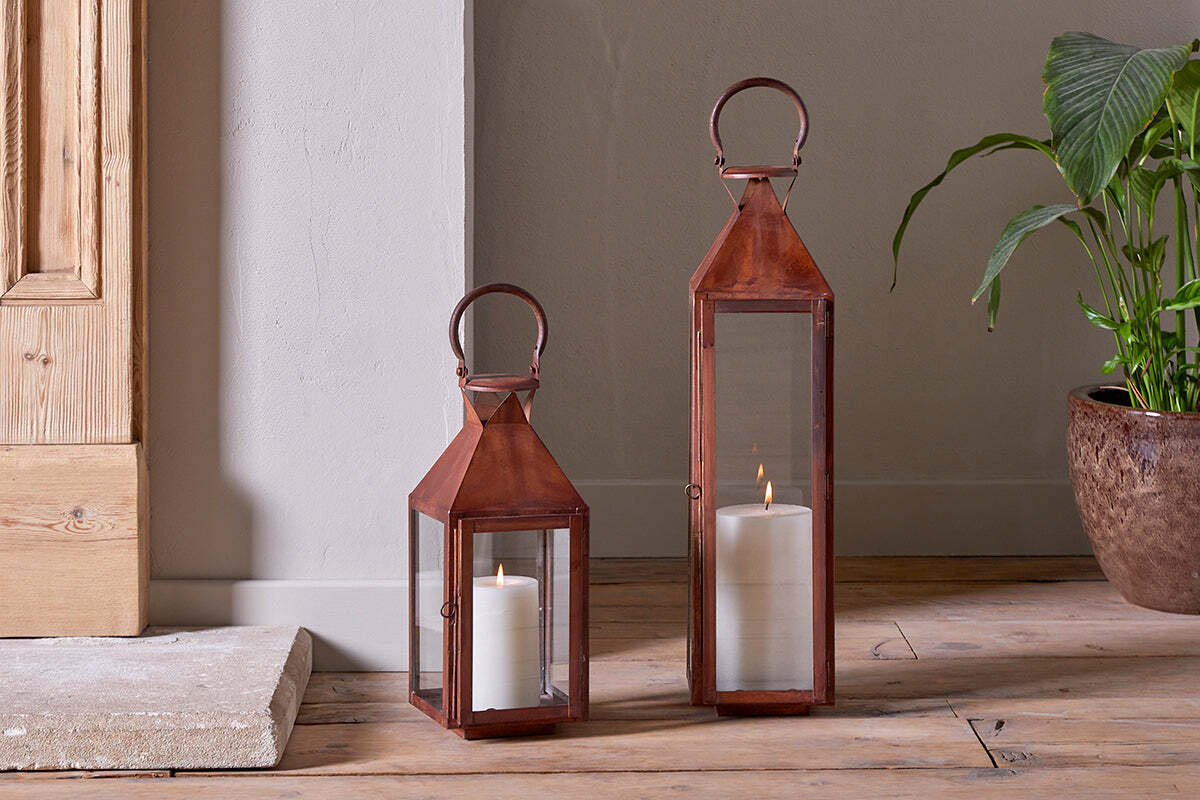 Two nkuku Isko Lanterns in a rustic rust finish, each with glass panels and a lit white pillar candle inside, sit on a wooden floor by a beige wall. A leafy potted plant, garden seating, and part of a wooden door appear in the background.