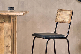 A wooden table with a mug stands beside the Iswa Leather & Cane Counter Chair by nkuku, featuring an aged black metal frame, black cushioned seat, and woven cane back, all set against a beige textured wall for a rustic, minimal look.