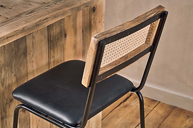 The nkuku Iswa Leather & Cane Counter Chair in Aged Black, featuring a woven cane back and black cushioned seat, sits by a rustic wooden table on a wooden floor, enhancing the space’s warm and minimalist appeal.