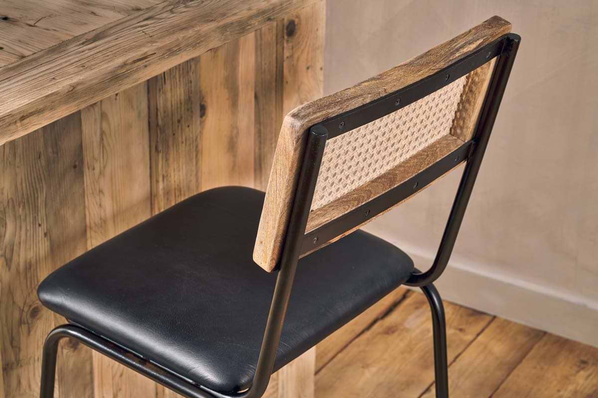 The nkuku Iswa Leather & Cane Counter Chair in Aged Black, featuring a woven cane back and black cushioned seat, sits by a rustic wooden table on a wooden floor, enhancing the space’s warm and minimalist appeal.