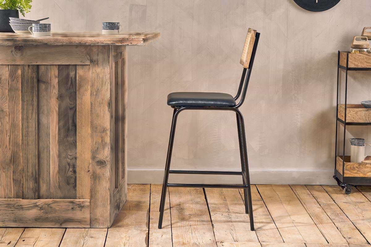The Iswa Leather & Cane Counter Chair by nkuku, featuring an aged black frame, black leather seat, and light cane backrest, is placed by a rustic wooden bar on wooden flooring with a small cart against a beige wall in the background.