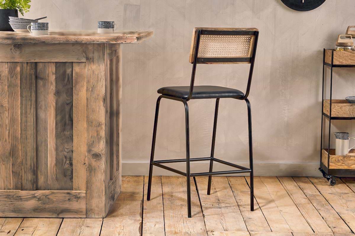The Iswa Leather & Cane Counter Chair by nkuku, featuring a woven cane back, black metal frame, and cushioned black leather seat in aged black, stands on a rustic wooden floor beside a wooden bar counter in a cozy, modern room.