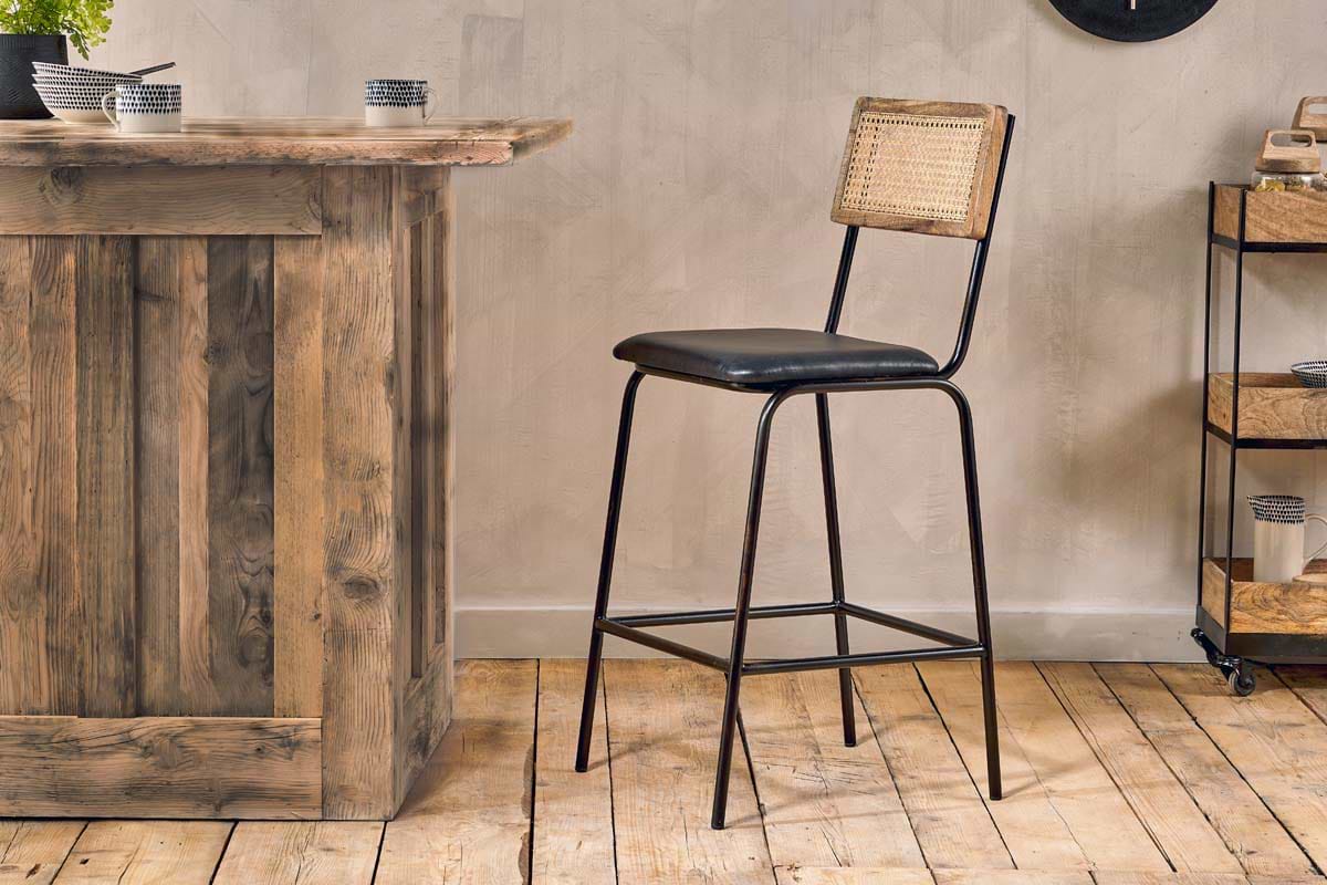A rustic kitchen scene with the nkuku Iswa Leather & Cane Counter Chair in Aged Black, featuring a black metal frame, woven cane back, and black leather seat beside a weathered wood bar on wooden floors with decor and a rolling cart in the background.