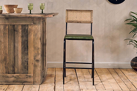 A wooden bar with dishes and glasses sits on a rustic wood floor. Next to it is the nkuku Iswa Leather & Cane Counter Chair in rich green, featuring cane backing, a green cushioned seat, and black metal legs—ideal for stylish kitchen island seating.