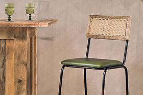 The Iswa Leather & Cane Counter Chair by nkuku features a woven cane back, black metal legs, and a rich green leather seat—ideal for stylish kitchen island seating beside rustic wooden bars and green glass goblets against beige walls.