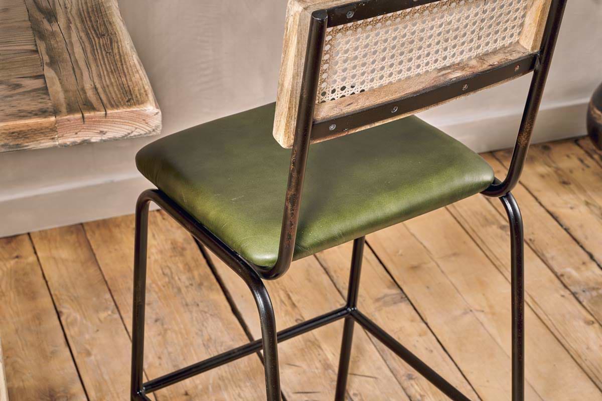 The Iswa Leather & Cane Counter Chair by nkuku features a rich green cushioned seat, metal frame, and woven rattan cane back—perfect for kitchen island seating against a beige wall and rustic table on a wooden floor.
