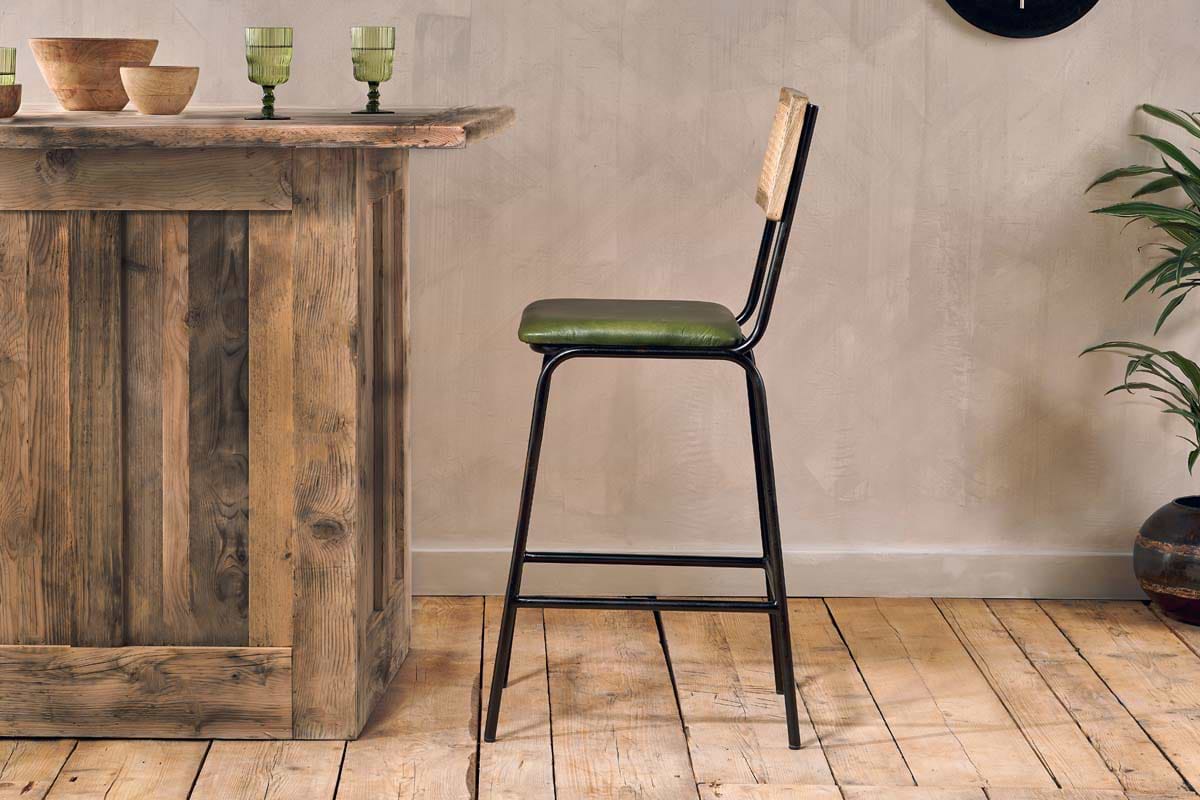 A rustic wooden bar sits on a light wood floor next to the nkuku Iswa Leather & Cane Counter Chair in rich green. Three green glasses, wooden bowls, and a leafy potted plant decorate the space against the beige wall.
