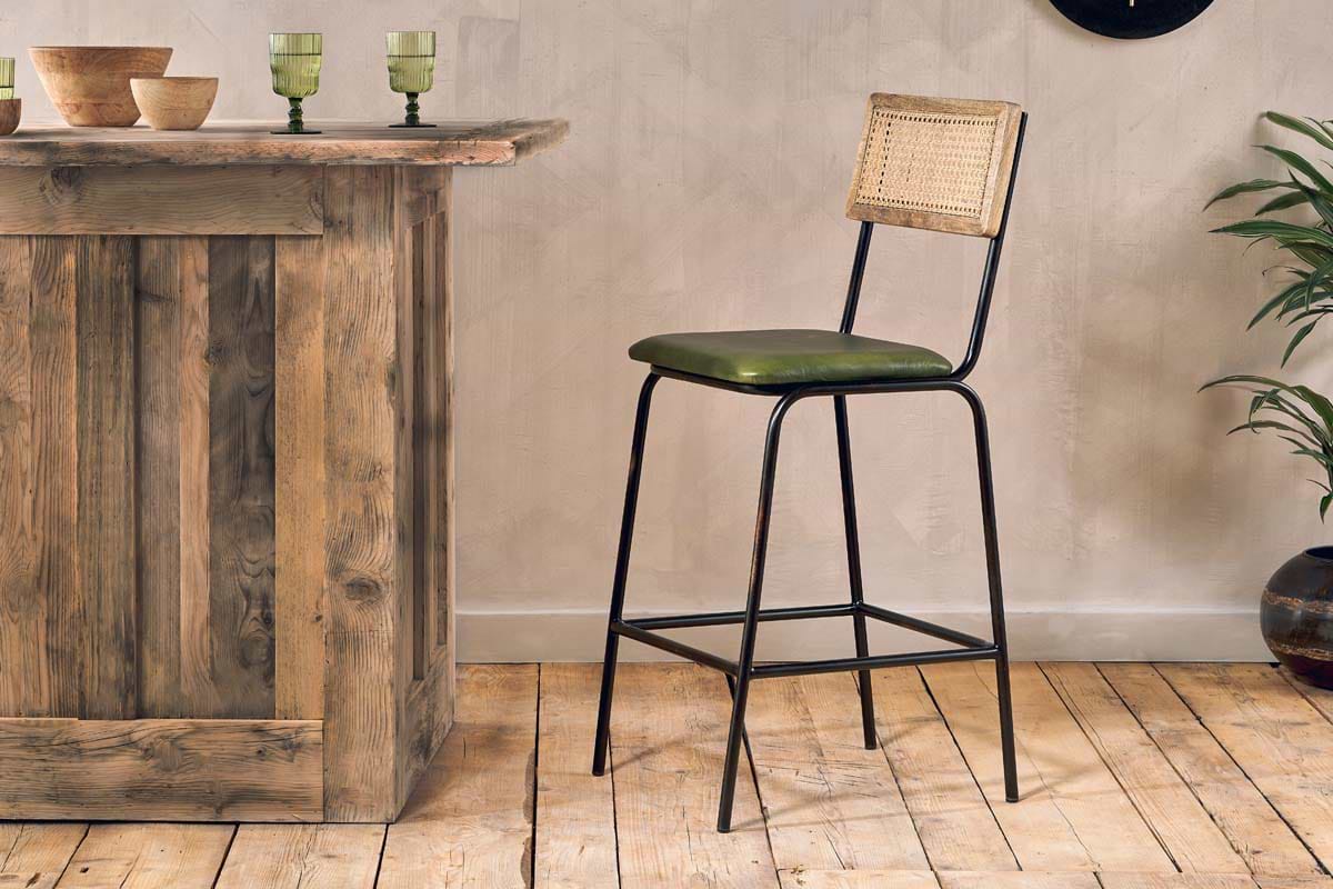 A rustic wooden kitchen island stands on a wood floor with the nkuku Iswa Leather & Cane Counter Chair in rich green, featuring black metal legs and a woven cane back. Green glassware and a potted plant complete the inviting space.