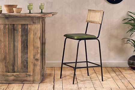 A rustic wooden kitchen island stands on a wood floor with the nkuku Iswa Leather & Cane Counter Chair in rich green, featuring black metal legs and a woven cane back. Green glassware and a potted plant complete the inviting space.