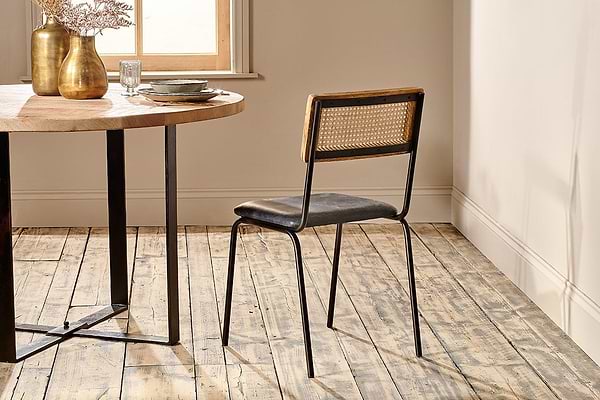 The Iswa Leather & Cane Dining Chair - Black by nkuku, featuring a mango wood frame and dark seat, sits beside a round wooden table with dishes and gold vases. The room has wooden floors, cream walls, and sunlight streaming in.