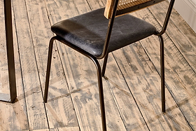 Close-up of the Iswa Leather & Cane Dining Chair - Black by nkuku, crafted with black goat leather and a mango wood frame, shown on distressed wooden floorboards.