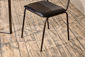 The nkuku Iswa Leather & Cane Dining Chair in black, featuring a slim metal frame and gently aged goat leather seat, is set on a distressed light wood floor with visible grain. A metal table leg appears on the image's left side.