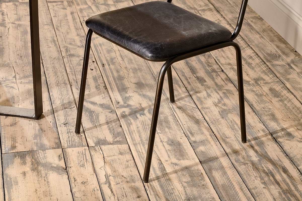 The nkuku Iswa Leather & Cane Dining Chair in black, featuring a slim metal frame and gently aged goat leather seat, is set on a distressed light wood floor with visible grain. A metal table leg appears on the image's left side.