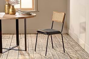 The Iswa Leather & Cane Dining Chair - Black by nkuku showcases a mango wood frame, black goat leather seat, and cane back, positioned beside a round wooden dining table with black metal legs on a rustic wooden floor.