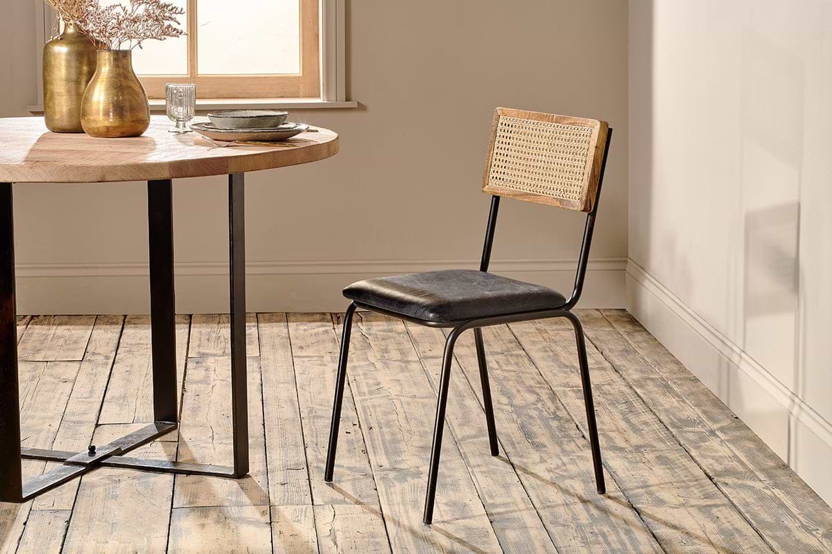 The Iswa Leather & Cane Dining Chair - Black by nkuku showcases a mango wood frame, black goat leather seat, and cane back, positioned beside a round wooden dining table with black metal legs on a rustic wooden floor.