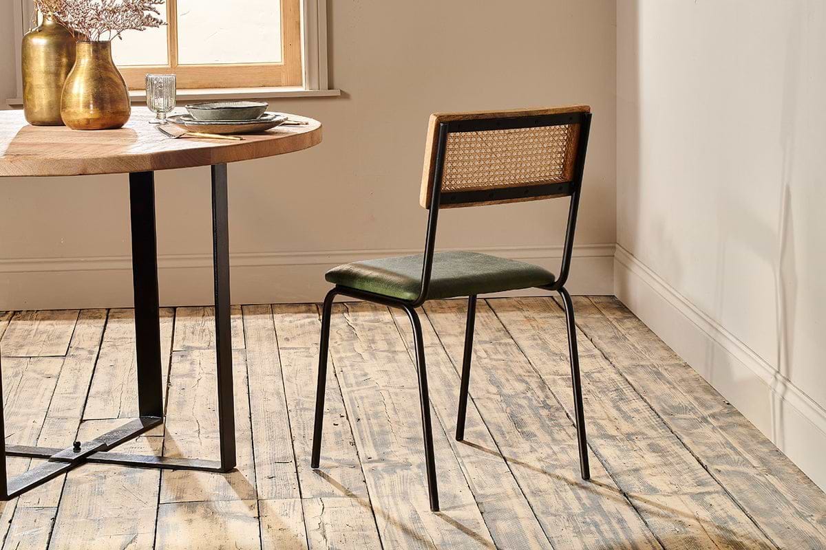 A dining room with a wooden table set for one, a gold vase, and the nkuku Iswa Leather & Cane Dining Chair in green. Sunlight streams through a window, illuminating rustic wooden floors and cream walls.