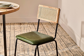 The nkuku Iswa Leather & Cane Dining Chair - Green, featuring an industrial black iron frame and light wood backrest, is placed on a rustic floor beside a round table. The room has beige walls and white baseboards.