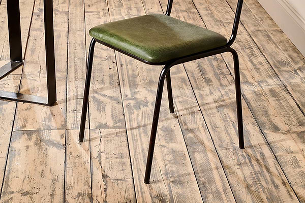 The Iswa Leather & Cane Dining Chair - Green by nkuku, featuring a green cushioned seat and black metal legs, is placed on a rustic wooden floor near an off-white wall and next to a black metal table.