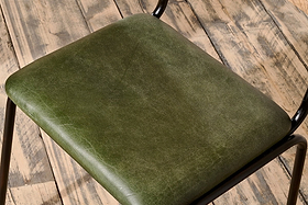 Close-up of the Iswa Leather & Cane Dining Chair in green by nkuku, featuring dark green goat leather with subtle creases, a black iron frame, and a cane seat, set on a rustic wooden floor.