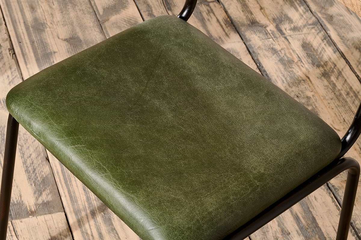 Close-up of the Iswa Leather & Cane Dining Chair in green by nkuku, featuring dark green goat leather with subtle creases, a black iron frame, and a cane seat, set on a rustic wooden floor.