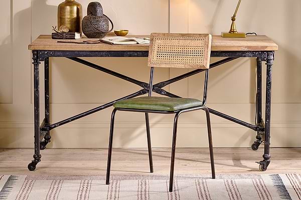 A rustic wooden desk with an industrial black iron frame and wheels is paired with the nkuku Iswa Leather & Cane Dining Chair in green. Decorative items sit atop the desk.