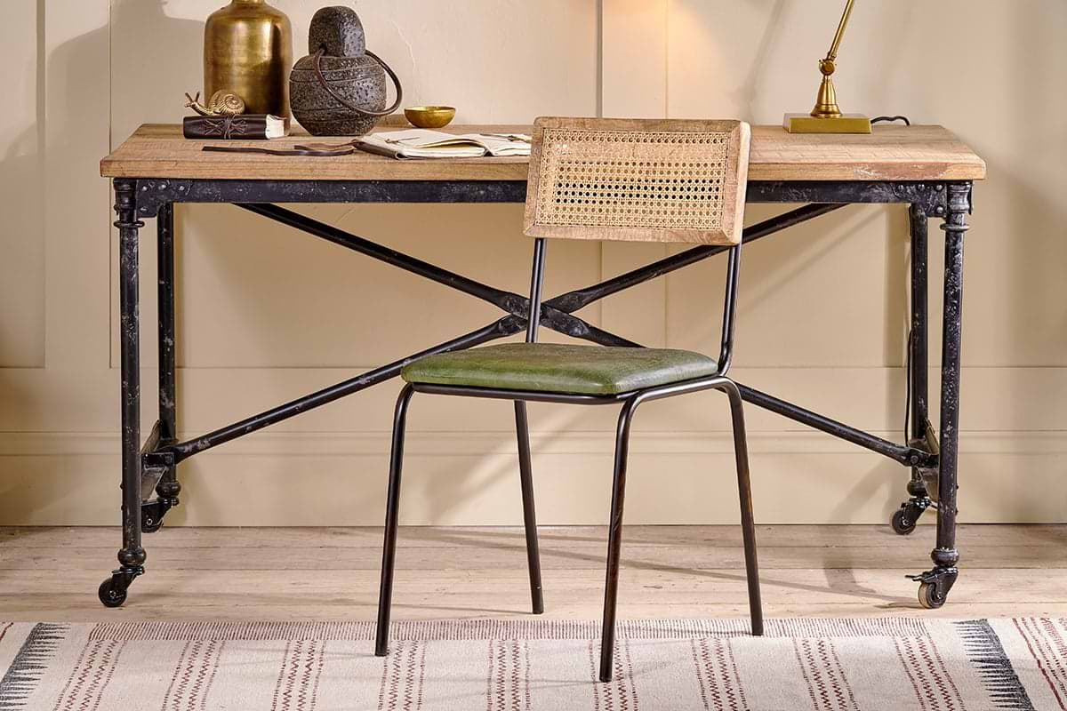 A rustic wooden desk with an industrial black iron frame and wheels is paired with the nkuku Iswa Leather & Cane Dining Chair in green. Decorative items sit atop the desk.