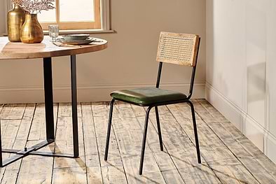 The rustic dining set features a round wooden table with an industrial black iron frame, paired with the nkuku Iswa Leather & Cane Dining Chair in green, which has a cane-backed wooden frame and a green upholstered seat.