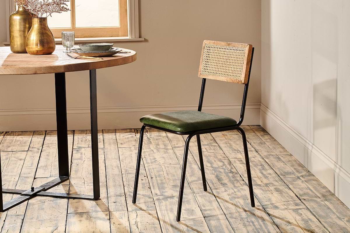 The rustic dining set features a round wooden table with an industrial black iron frame, paired with the nkuku Iswa Leather & Cane Dining Chair in green, which has a cane-backed wooden frame and a green upholstered seat.