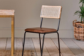The nkuku Iswa Leather & Cane Dining Chair - Tan, featuring a cane back and brown leather seat, is placed on a wooden floor next to a dining table.