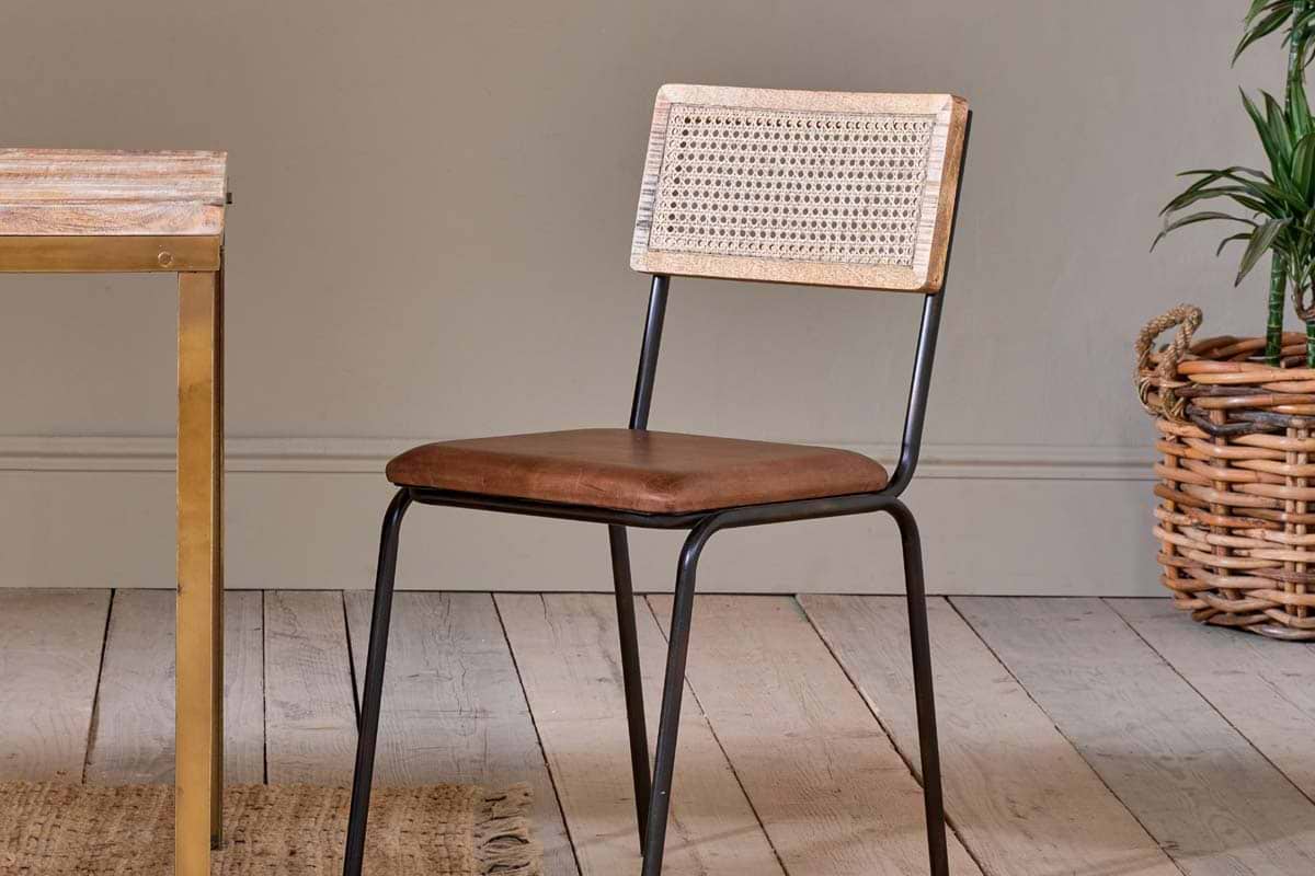 The nkuku Iswa Leather & Cane Dining Chair - Tan, featuring a cane back and brown leather seat, is placed on a wooden floor next to a dining table.