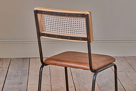 The Iswa Leather & Cane Dining Chair - Tan by nkuku features a woven cane back, tan leather seat, and dark metal frame. Ideal for kitchen dining tables, it enhances any room with its stylish design against a neutral wall and light wooden floor.