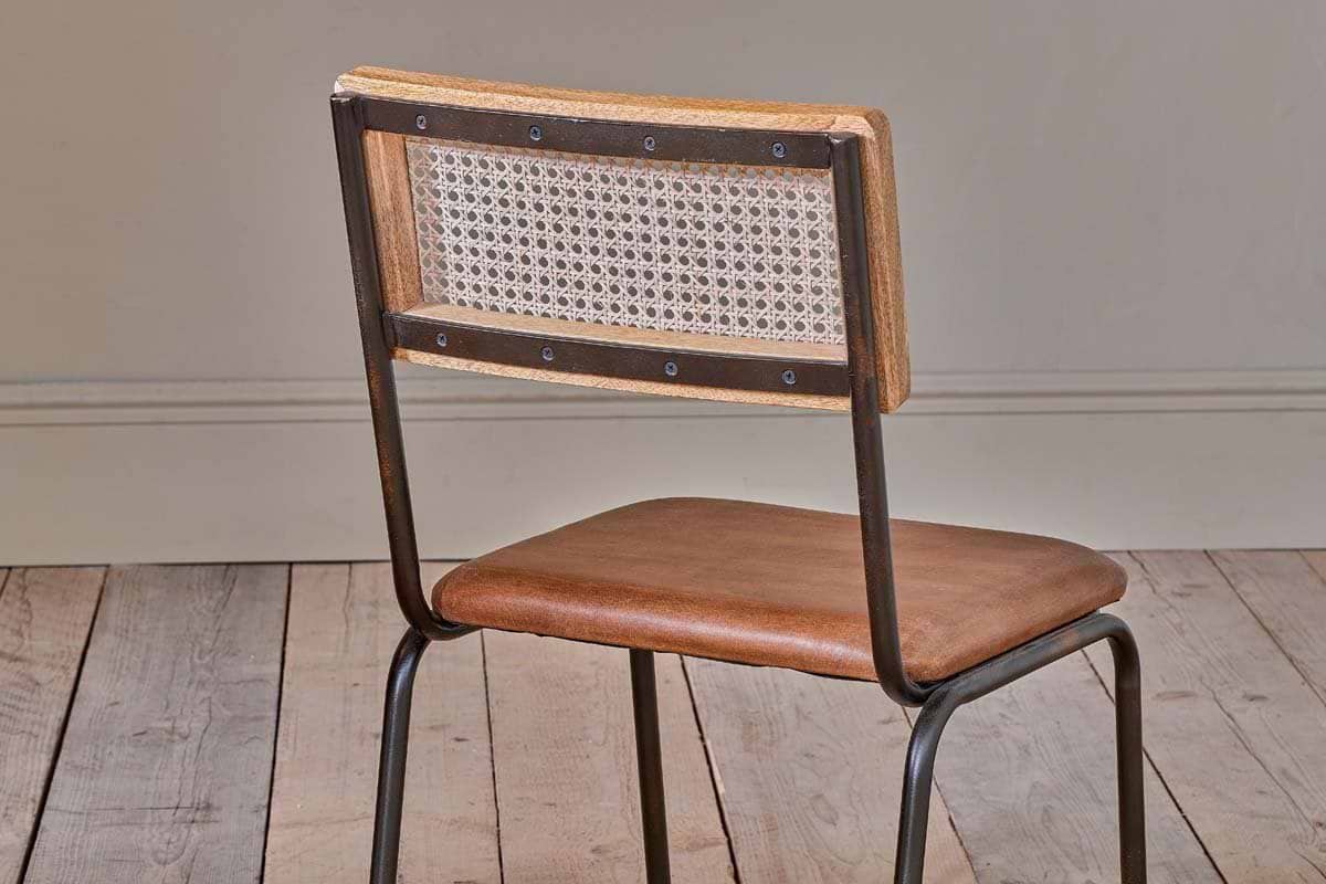 The Iswa Leather & Cane Dining Chair - Tan by nkuku features a woven cane back, tan leather seat, and dark metal frame. Ideal for kitchen dining tables, it enhances any room with its stylish design against a neutral wall and light wooden floor.