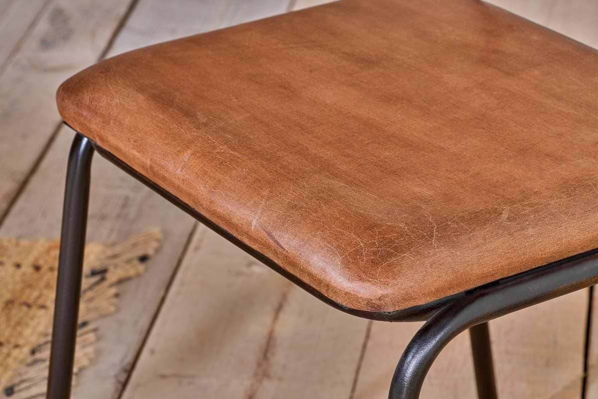 Close-up of the nkuku Iswa Leather & Cane Dining Chair in tan, featuring square-shaped, light brown leather upholstery with rounded edges on black metal legs. Wooden floorboards and a woven rug appear in the background.