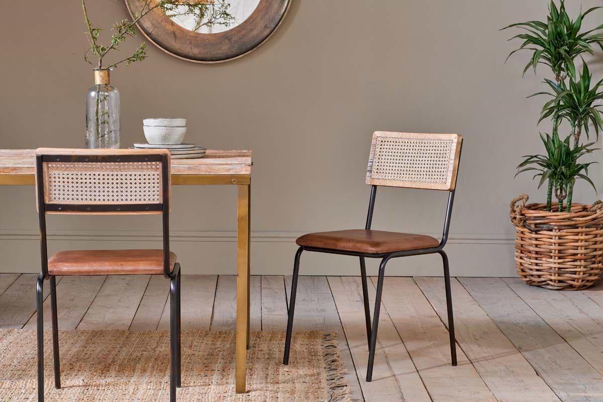 Two nkuku Iswa Leather & Cane Dining Chairs in tan sit at a mango wood table. A plant in a woven basket stands to the right.