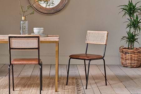 Two nkuku Iswa Leather & Cane Dining Chairs in tan sit at a mango wood table. A plant in a woven basket stands to the right.
