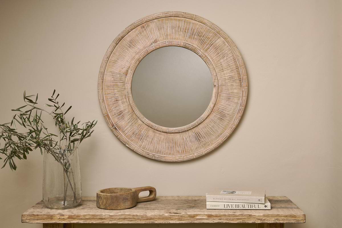 The nkuku Itwa Mango Wood Carved Mirror in natural hangs on a beige wall above a wooden table styled with a glass vase of olive branches, a handled wooden bowl, and stacked books for a simple, minimalist look.