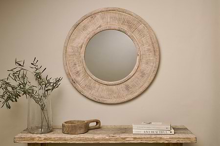 The nkuku Itwa Mango Wood Carved Mirror in natural hangs on a beige wall above a wooden table styled with a glass vase of olive branches, a handled wooden bowl, and stacked books for a simple, minimalist look.