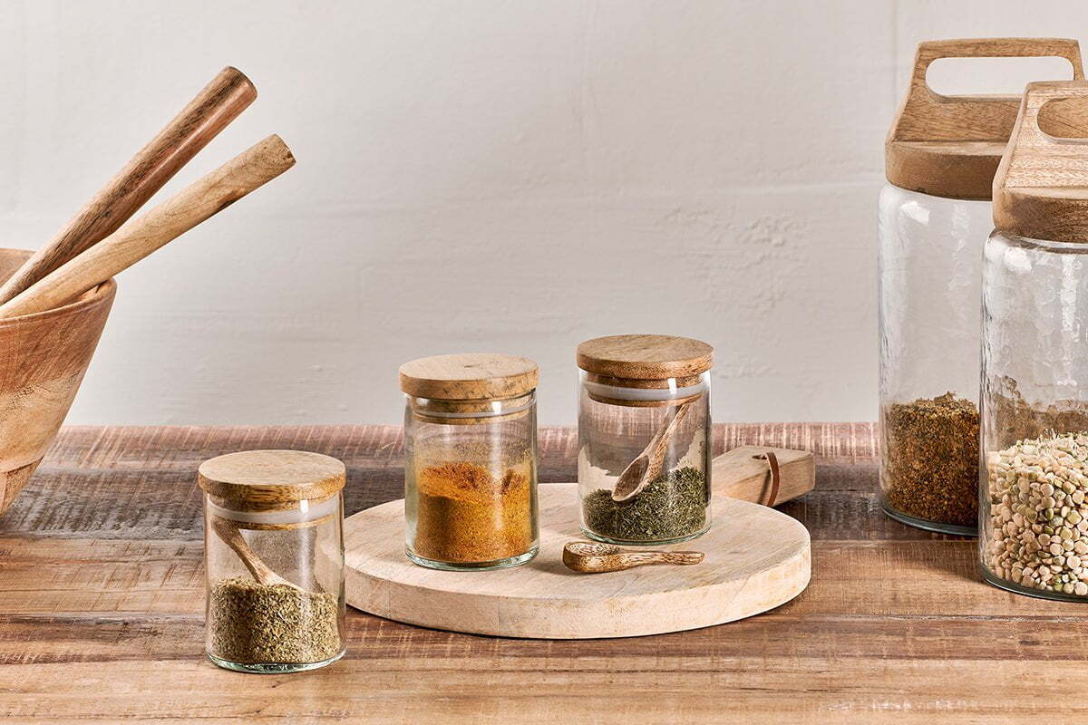The nkuku Izaan Spice Jar set features three small clear jars with mango wood lids and spoons, elegantly arranged on a round mango wood board—perfect for adding rustic charm to your kitchen.