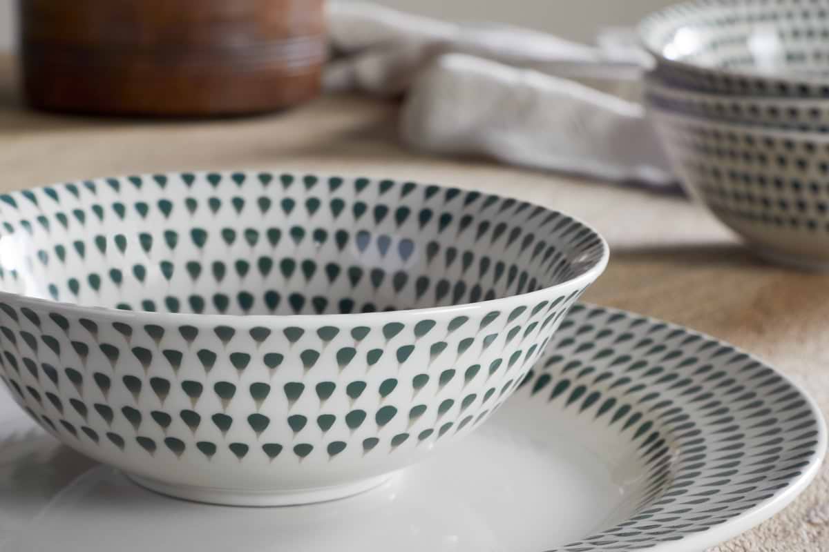 A close-up of the nkuku Juniper Drop Cereal Bowl - Cream & Juniper, with its green drop pattern, sits on a matching plate. In the background, a similar bowl and textured cloth complete this inviting dining scene.