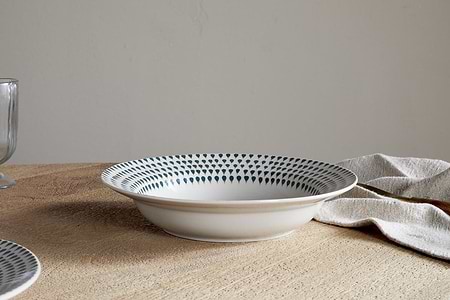 The Juniper Drop Pasta Bowl - Cream & Juniper by nkuku is a contemporary white ceramic bowl with a blue dotted rim, shown on a wooden table beside a beige napkin, glass, and partial plate.