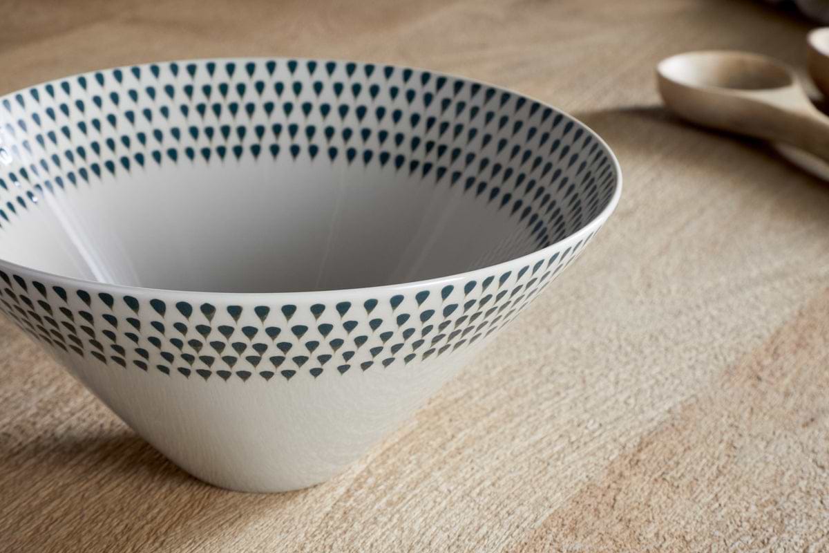 The nkuku Juniper Drop Serving Bowl in Cream & Juniper (Medium) features a stylish pattern of dark blue triangles around the rim and sits on a light wooden surface, with wooden utensils blurred in the background, making it an elegant table centrepiece.