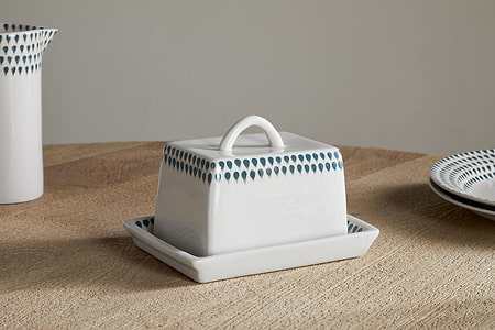 The nkuku Juniper Drop Butter Dish - Cream & Juniper features a white ceramic base and lid with a blue teardrop pattern, displayed on a light wooden table next to matching plates.