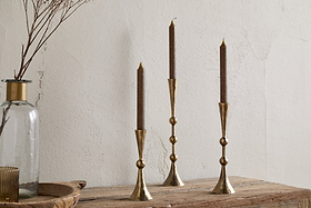 Three nkuku Javanee Candle Holders in antique brass with brown taper candles are arranged on a rustic wooden surface. Nearby, a clear glass vase of dried branches adds to the sculptural scene against a textured light wall.