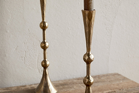 Two nkuku Javanee Candle Holders in antique brass, featuring a geometric hammered texture and spherical accents, stand on a rustic wooden surface with one partially visible on the left against a white textured wall.