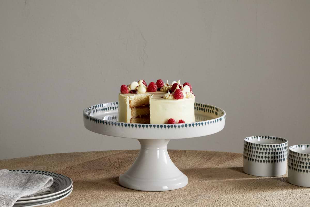 A small frosted cake with raspberries and cream sits on the nkuku Juniper Drop Ceramic Stand - Cream & Juniper, placed on a wooden table beside green drop-patterned cups and a folded napkin. One slice has been removed.