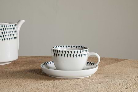 A nkuku Juniper Drop Cup & Saucer - Cream & Juniper, featuring a dark blue teardrop design, rests on a wooden table. The matching teapot from the set is partially visible on the left against a plain background.