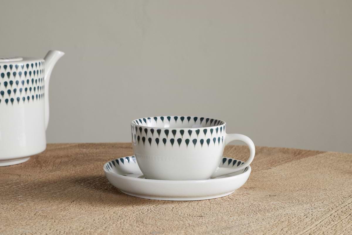 A nkuku Juniper Drop Cup & Saucer - Cream & Juniper, featuring a dark blue teardrop design, rests on a wooden table. The matching teapot from the set is partially visible on the left against a plain background.