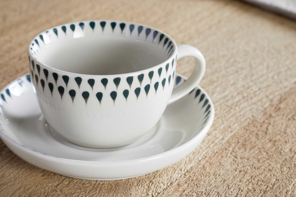 A nkuku Juniper Drop Cup & Saucer - Cream & Juniper, featuring a blue-green drop rim pattern, rests on its matching saucer atop a textured wooden surface.
