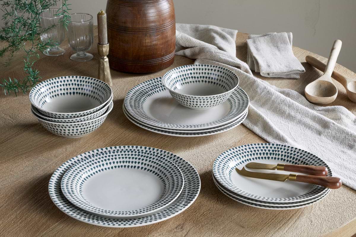 A wooden table is set with the nkuku Juniper Drop Dinnerware Set - Cream & Juniper (Set of 12) ceramic plates and bowls, a wooden container, two glasses, a candle, cloth napkins, wooden utensils, and a sprig of juniper green as decor.