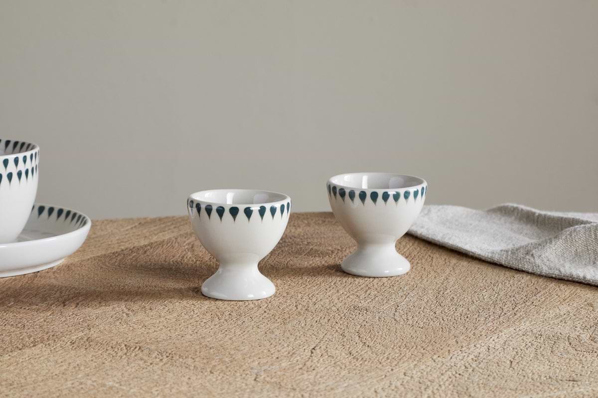 The Juniper Drop Egg Cups - Cream & Juniper by nkuku (set of 2) with blue teardrop patterns sit on a textured brown table next to a folded beige cloth. Matching plate and bowl partially visible on the left.